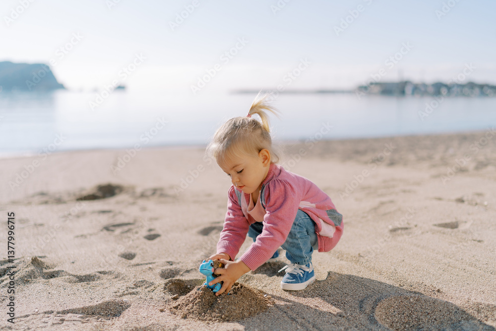 Little girl squats on the beach and pours sand into a toy dinosaur Little girl squats on the beach and pours sand into a toy dinosaur