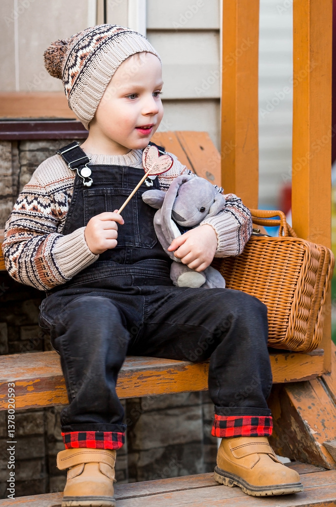 Childhood. A little boy in stylish clothes is sitting on a wooden porch with a lollipop and a soft toy outside the house in the fresh air. Happy childhood.