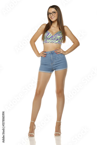 Smiling young woman with glasses wearing colorful patterned crop top and high-waisted denim shorts with hands on hips, posing in high heels