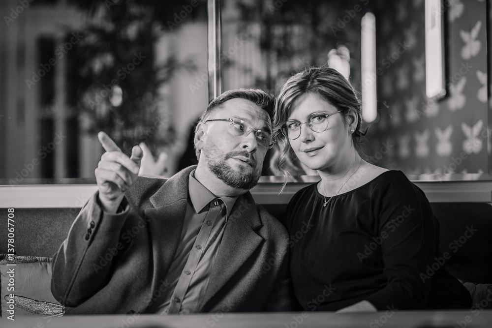 Obraz premium A man and woman sit closely on a couch in a stylish indoor setting. The man gestures while talking, and the woman looks at the camera. Black and white photo..