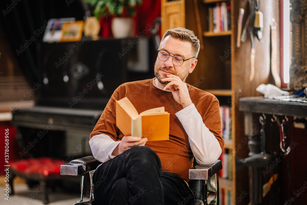 Fototapeta premium A man with glasses and a beard reads a book while seated in a cozy room. He appears calm and focused, holding his chin thoughtfully..