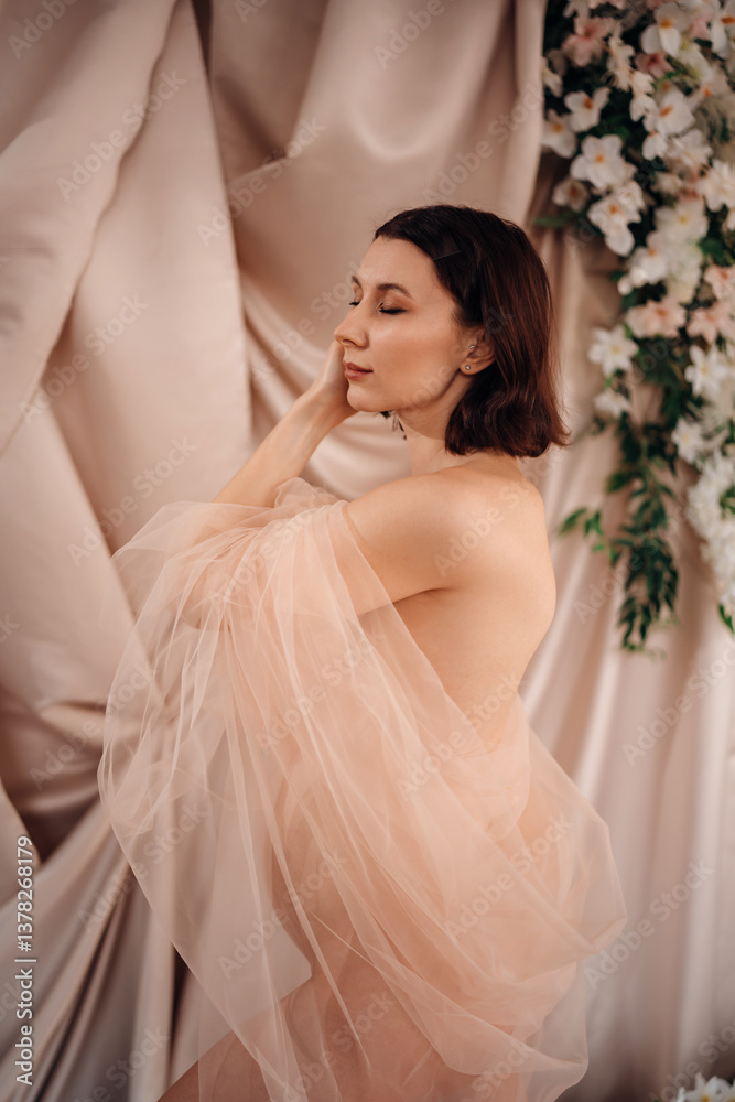 Fototapeta premium Pregnant young woman in soft fabric poses gracefully against a floral backdrop