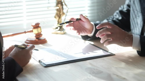 Two men are sitting at a desk with a pen and a piece of paper. One of the men is holding a pen and pointing at the paper. The other man is looking at the paper and the pen