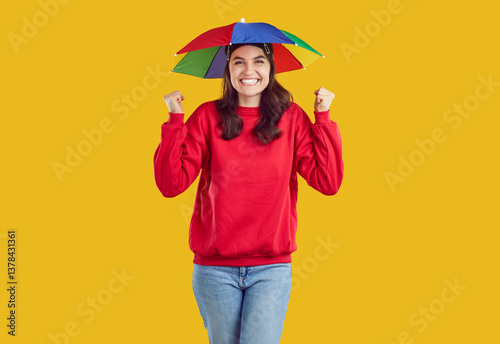 Happy woman with colorful head umbrella sincerely rejoices, isolated on orange background. Woman with original rainbow umbrella on her head from sun or rain is joyfully clenching her fists, rejoicing.