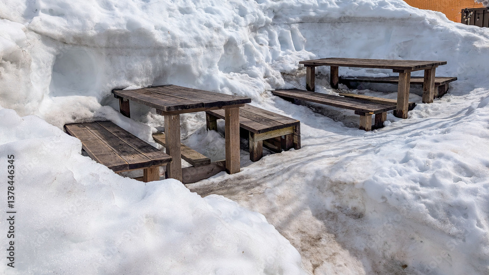 Wooden picnic benches surrounded by deep snow on a sunny day at a ski resort