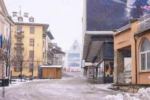 Obraz na płótnie A snowy street in Cortina d'Ampezzo, Italy, featuring Alpine style buildings, a church tower, and a billboard for the Winter Olympics 2026. w salonie
