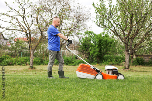 Man in protective gloves, gardener is mowing bright lush green grass lawn using modern gasoline cordless lawnmower at own backyard. Seasonal landscaping design work. Gardening work tools.