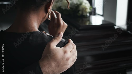 Close-up over the shoulder shot of tearful African American woman comforted by unrecognizable male hand, standing in front of coffin at funeral