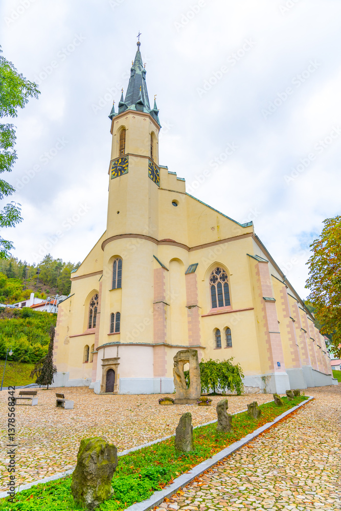 Naklejka premium The Church of St. Joachim stands elegantly in Jachymov, Czechia, showcasing its architectural beauty amidst lush greenery and cloudy skies. Visitors admire the historic structure.