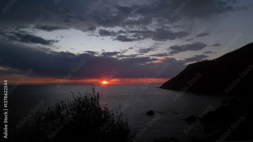 beautiful scenic epic drone flight over the sea at sunset with the red sun setting into the sea on the horizon with a colorful sky full of gray clouds in Framura Italy Liguria landscape