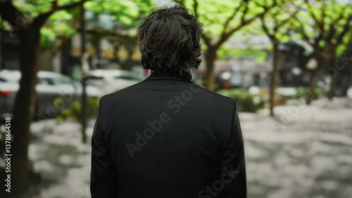 Man in suit standing outdoors in a city street with green trees lining the background creating a natural urban environment.