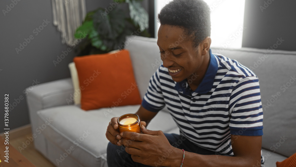 Fototapeta premium Young man relaxing indoors smiling and holding a lit candle, sitting on a comfortable sofa in a stylish living room apartment with warm decor and natural lighting.