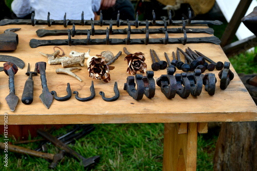 A close up on a number of blacksmith tools and creations, including horseshoes, hooks, pikes, knives, and hammers displayed on a wooden table during a medieval fair in Poland