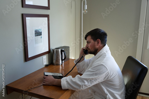 Handsome caucasian man in the hotel room wearing white bathrobe, picked the phone to call guest service attendant to make an room service order