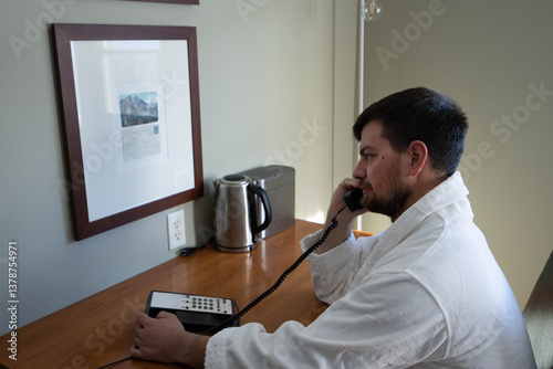 Handsome caucasian man in the hotel room wearing white bathrobe, picked the phone to call guest service attendant to make an room service order