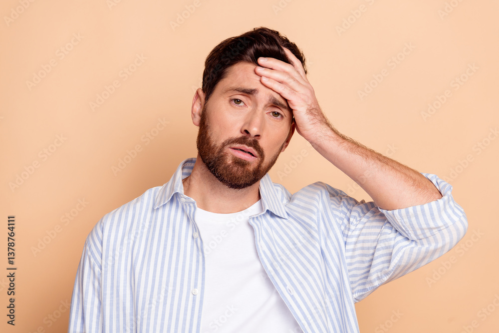 Young man with a hand on his forehead looking thoughtful in casual striped shirt against a beige background Young man with a hand on his forehead looking thoughtful in casual striped shirt against a beige background