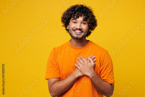 Foto Young smiling thankful cheerful happy Indian man he wearing orange t-shirt casual clothes put folded hands on heart look camera isolated on plain yellow background studio portrait