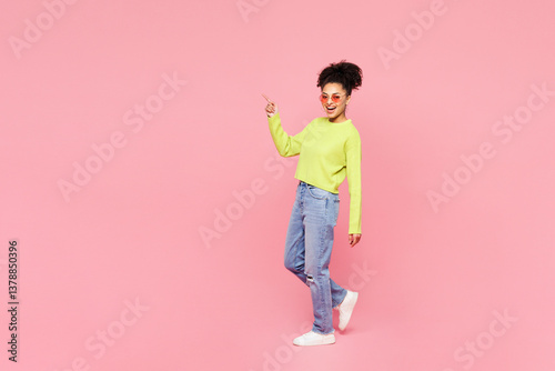 Full body young woman of African American ethnicity she wear green sweater casual clothes walk go point index finger aside on area mock up isolated on plain pastel pink background. Lifestyle concept. photo