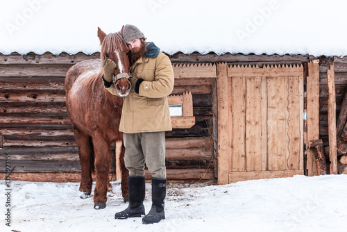 A man with a beard stands hugging a horse and looks at the camera, portrait, winter, snow, village, peasant, farm, wooden house.