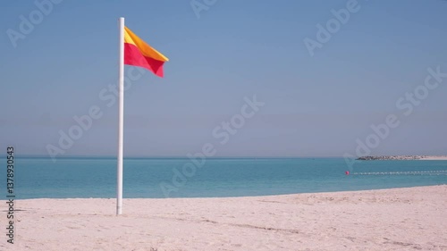 Yellow and red beach safety flag stands prominently on the sandy shoreline under a clear blue sky. The calm sea stretches into the horizon, symbolizing a serene beach day.