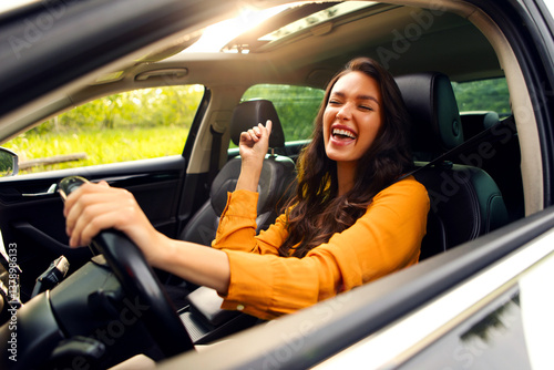 Overjoyed female driver enjoying auto ride, listening to music and singing while holding one hand on steering wheel, riding on sunny day