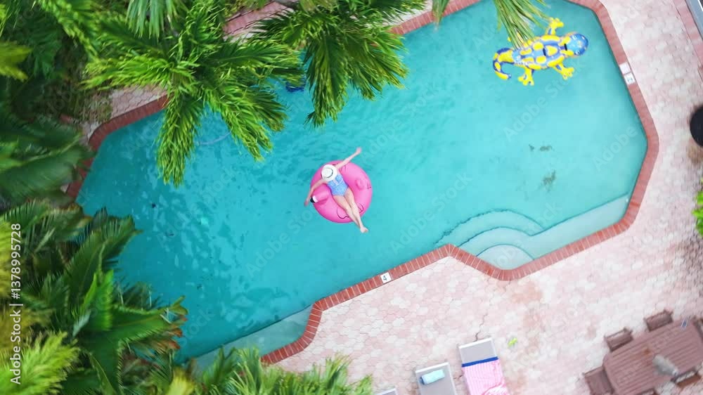 Top view of vibrant pool surrounded by palm trees, with elegant woman relaxing on pink flamingo float. Aerial swimming pool in tropical garden Miami, Florida. Woman in pool during summer vacation 4K