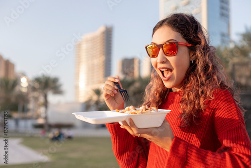 Obraz na płótnie Happy young woman enjoying street food in a city park, holding a takeaway box and savoring delicious meal w salonie