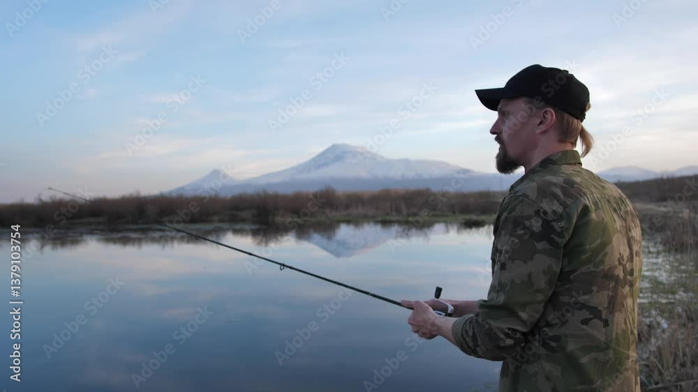 Young male fisherman uses spinning rod swinging and casting bait into water started to rotate fishing reel. Evening fishing near beautiful mountains with snowy peaks. Ararat Young male fisherman uses spinning rod swinging and casting bait into water started to rotate fishing reel. Evening fishing near beautiful mountains with snowy peaks. Ararat