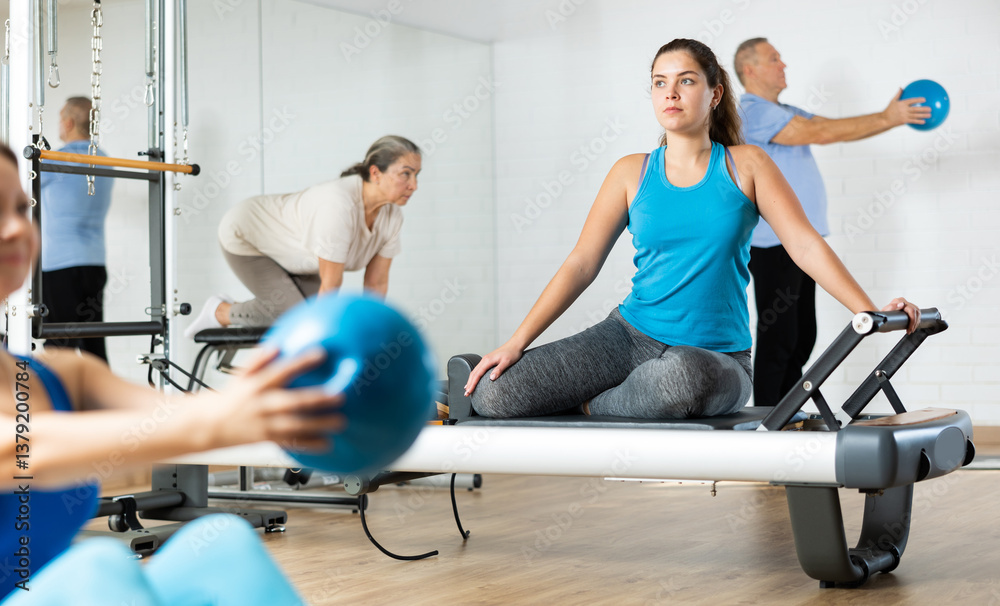 Positive concentrated young girl practicing pilates stretching exercises on reformer at gym..