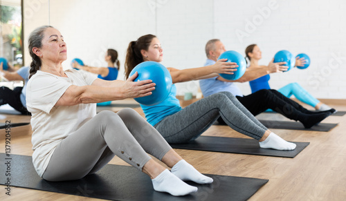 Sedulous old-aged woman doing pilates exercises with ball in gym area during workout session. Persons engaging in pilates in fitness room