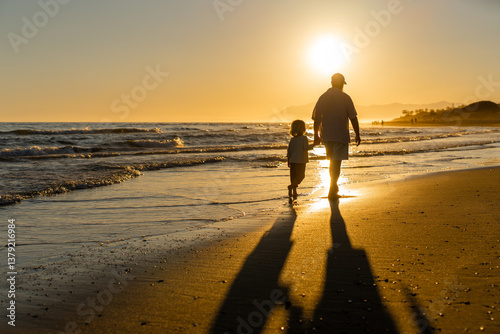 Grandfather and grandson walking barefoot along the beach at sunset, holding hands and casting long shadows in golden evening light by the sea.