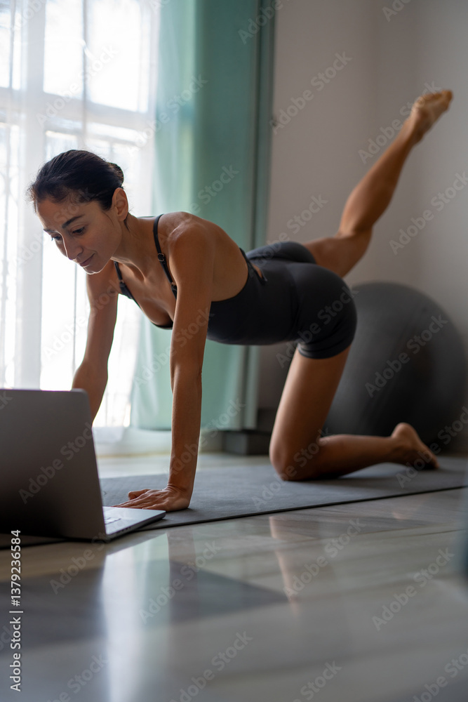 Fototapeta premium Focused Woman Doing Home Workout with Laptop Guidance on Yoga Mat