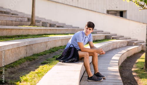 Teenage boy student 17 years old with a backpack sits on the wooden steps outside the college