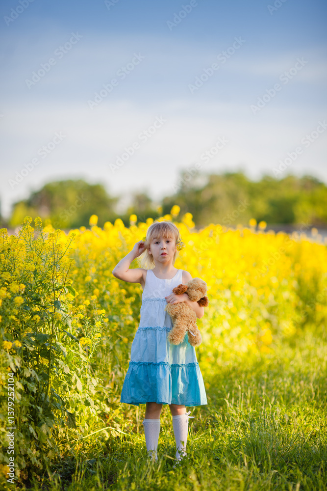 Fototapeta premium Little girl holding flower and teddy bear in blooming yellow meadow