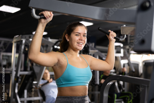 Cheerful sporty girl in blue top working out back muscles in modern gym, performing lat pull-down on lever machine. Physical activity and fitness concept