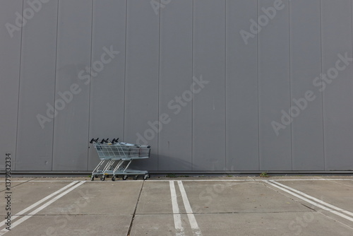 Minimalist image of three shopping carts aligned against a large gray industrial wall on an empty concrete parking lot. Urban scene with clean lines, negative space, and modern geometry