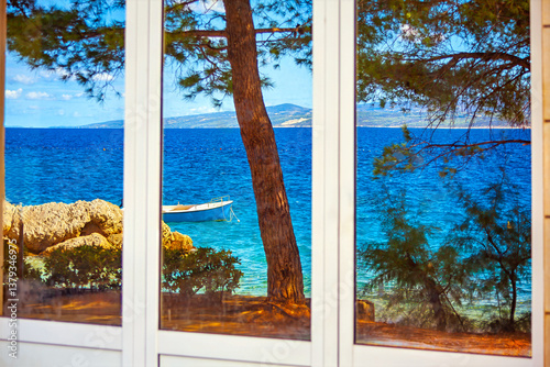 Glass window reflects stunning seaside view with small boat floating on turquoise water, framed by lush greenery and rocky shore. Serene coastal scene captures essence of relaxation and natural beauty