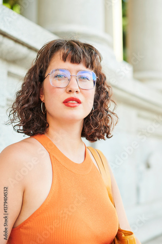 Real beauty of a young tourist woman with wavy hair, glasses, and an orange summer dress relaxing during a tour of a European city. Body acceptance concept.