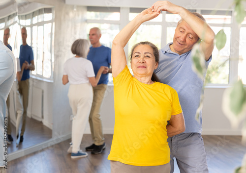 Positive senior man dancing playful Latin dance bachata with female partner during group class in choreography studio. Active lifestyle of older generation