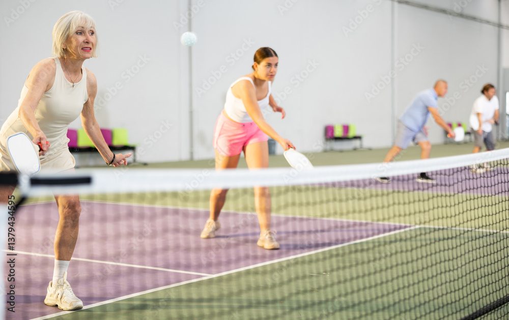Portrait of sporty senior woman playing doubles pickleball with young female partner on indoor court, ready to hit ball. Health and active lifestyle concept