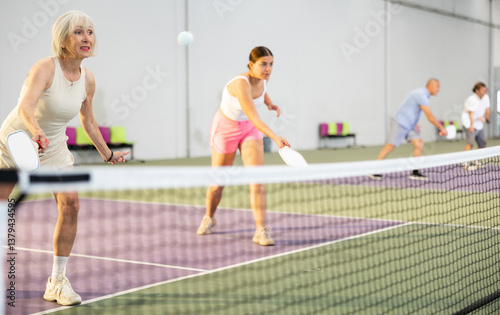 Obraz na płótnie Portrait of sporty senior woman playing doubles pickleball with young female partner on indoor court, ready to hit ball. Health and active lifestyle concept w salonie