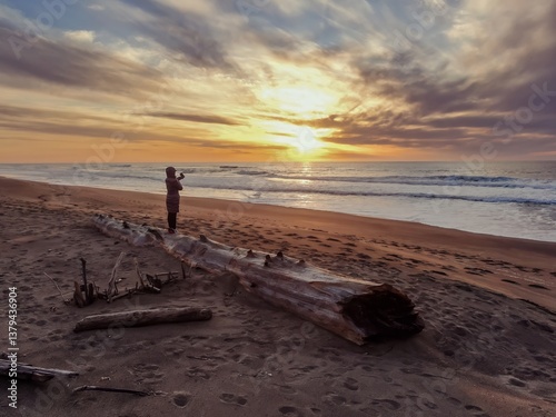 A woman stands on driftwood at a beach in Point Reyes Beach, Inverness, California, USA, taking a photo of the sunset over the ocean. The golden light reflects on the water and sand.