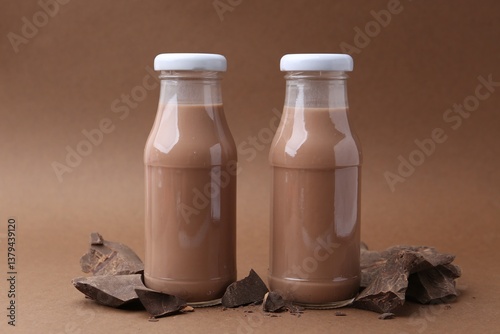 Tasty chocolate milk in glass bottles and pieces of chocolate on brown background, closeup
