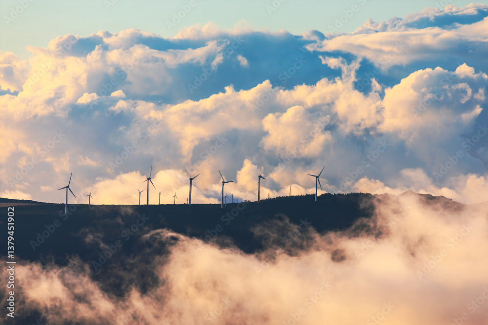 Wind turbines stands tall on mountain ridge, emerging through mist under dramatic sky filled with billowing clouds. Contrast of nature and renewable energy symbolizes sustainability and progress