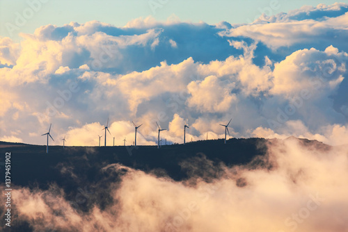 Obraz na płótnie Wind turbines stands tall on mountain ridge, emerging through mist under dramatic sky filled with billowing clouds. Contrast of nature and renewable energy symbolizes sustainability and progress w salonie