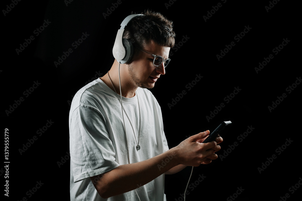 Naklejka premium Young man in white t-shirt and headphones looking at smartphone, listening to music or podcast. Isolated on black background. Modern digital lifestyle.