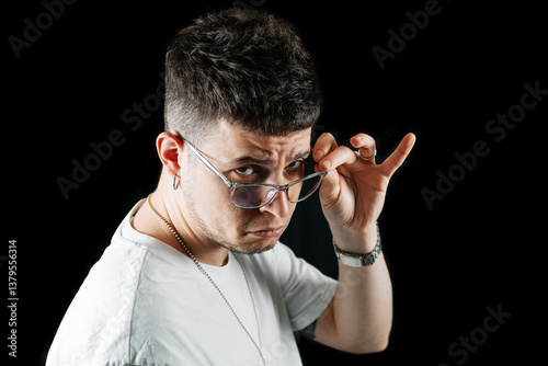 Canvas Print Confident young man in white t-shirt and transparent glasses lifting eyewear with ironic expression on face, isolated on black background