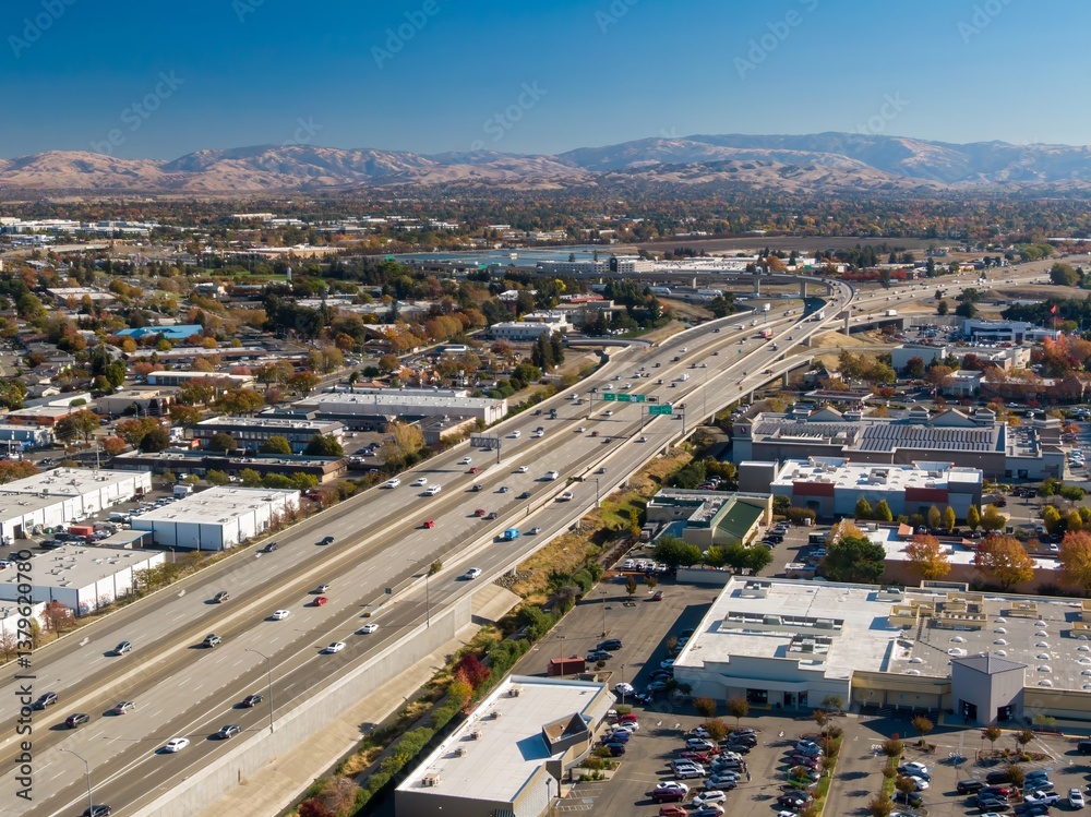 Fototapeta premium Aerial view of a busy Interstate 680 with cars moving through a suburban area with mountains in the background, Dublin, California, USA. Illustrates transportation and urban planning.