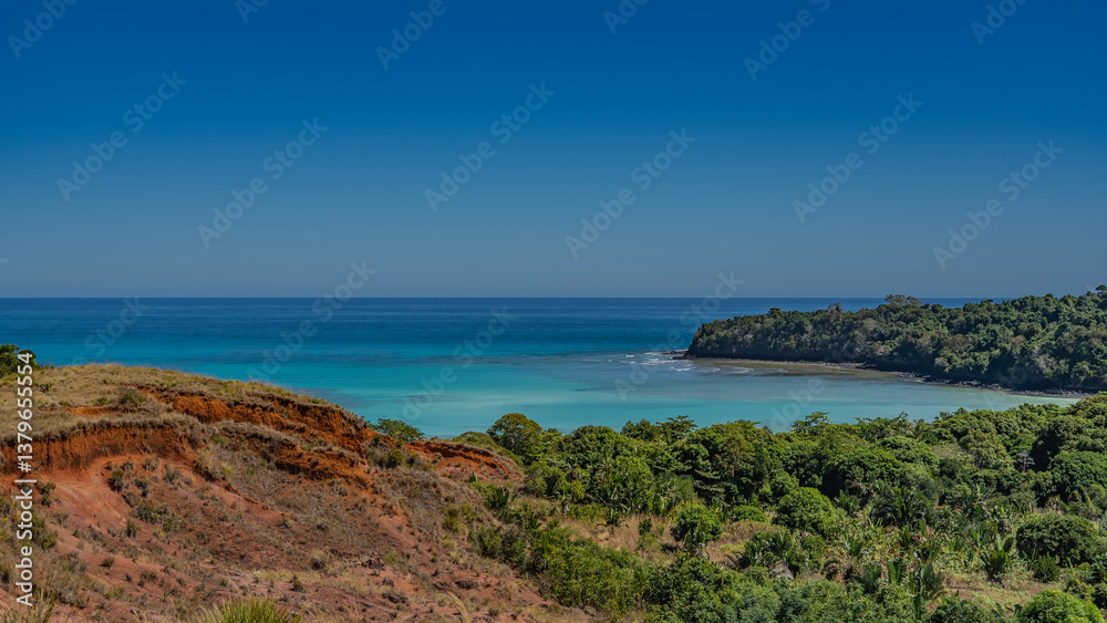 Beautiful serene seascape. A calm turquoise ocean, waves foaming at the shore. Lush green vegetation and a red-soil hill in the foreground. Clear blue sky. Copy space. Madagascar. Nosy Iranja Beautiful serene seascape. A calm turquoise ocean, waves foaming at the shore. Lush green vegetation and a red-soil hill in the foreground. Clear blue sky. Copy space. Madagascar. Nosy Iranja