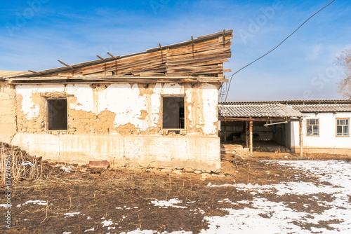 This image captures a mud-brick building with a partially collapsed wooden roof and broken windows. The house sits in a rural setting with patchy snow and dry ground highlighting the harsh environment